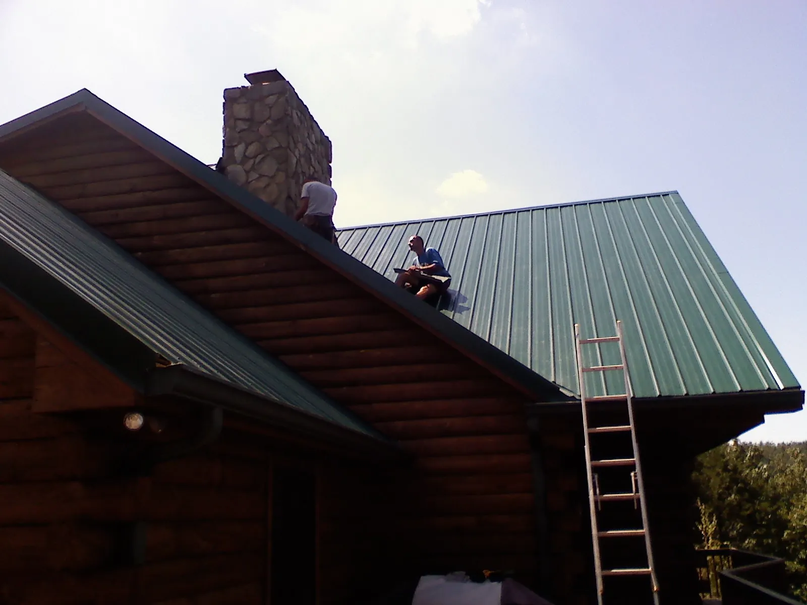 Roofers installing green metal panels beside a stone chimney on a log home in Orwell